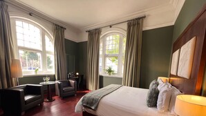 A hotel room with a large bed, a desk, a chair, and two windows with curtains. - Morgans Hotel (Swansea)