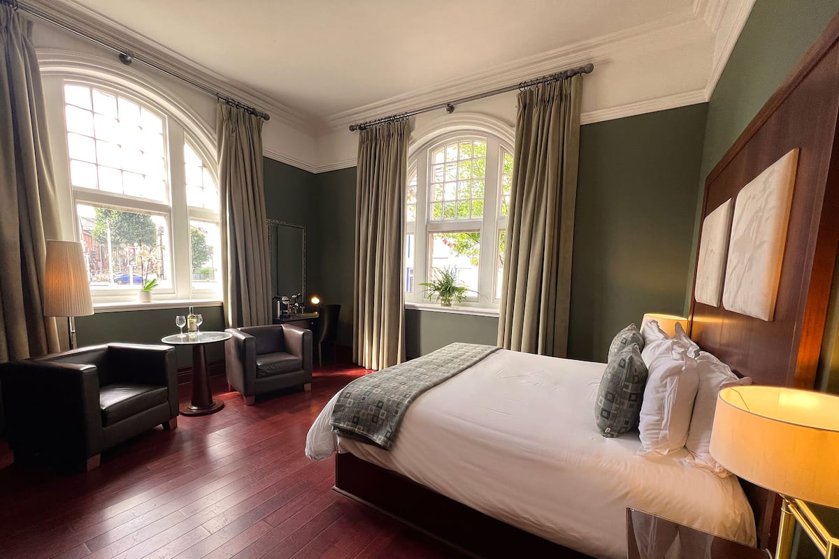 A hotel room with a large bed, a desk, a chair, and two windows with curtains.