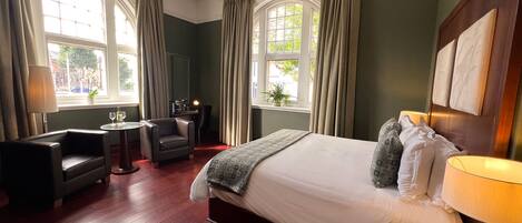 A hotel room with a large bed, a desk, a chair, and two windows with curtains.