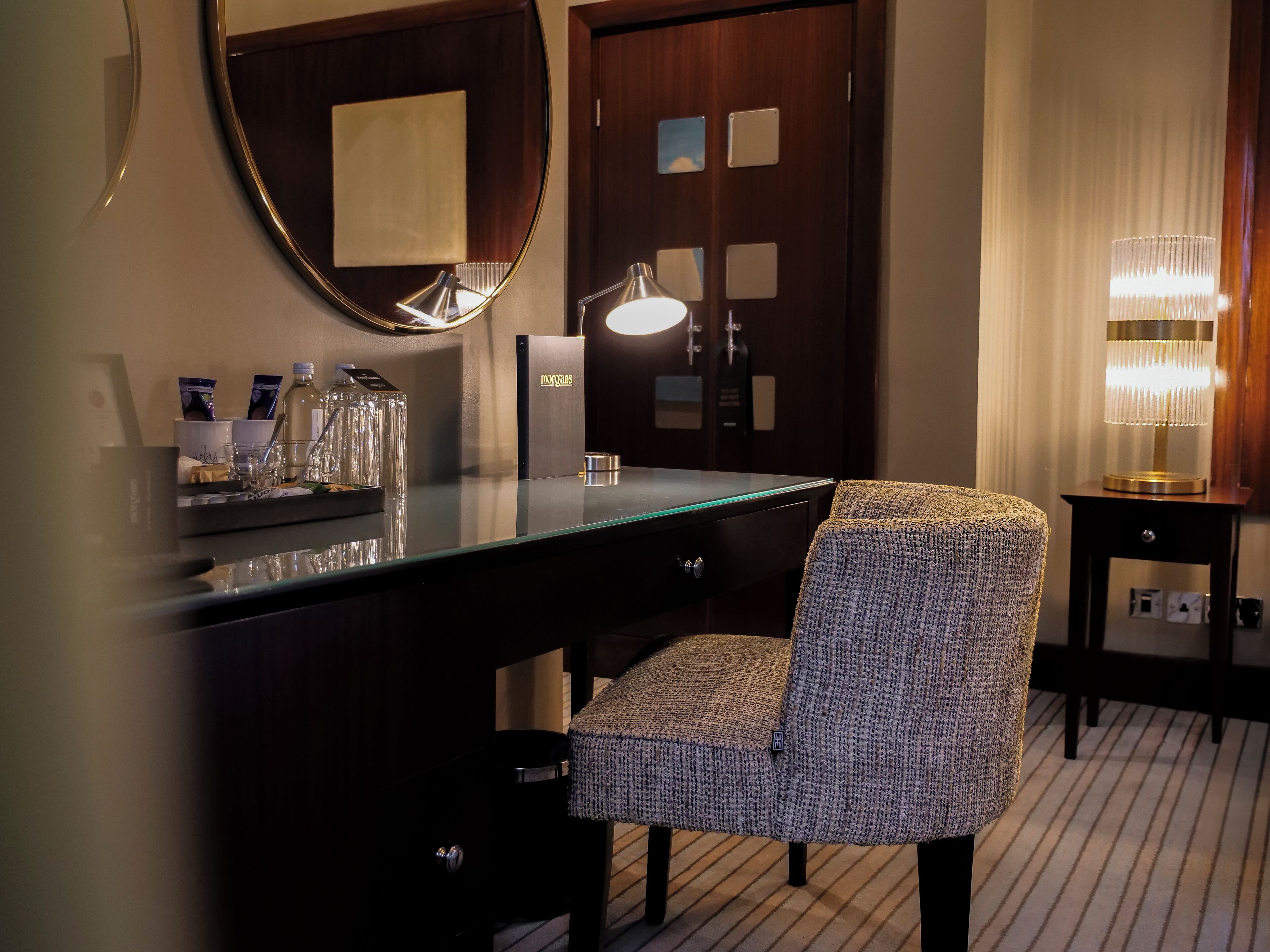 A hotel room with a dark wooden desk, a round mirror, a lamp, and a patterned chair.