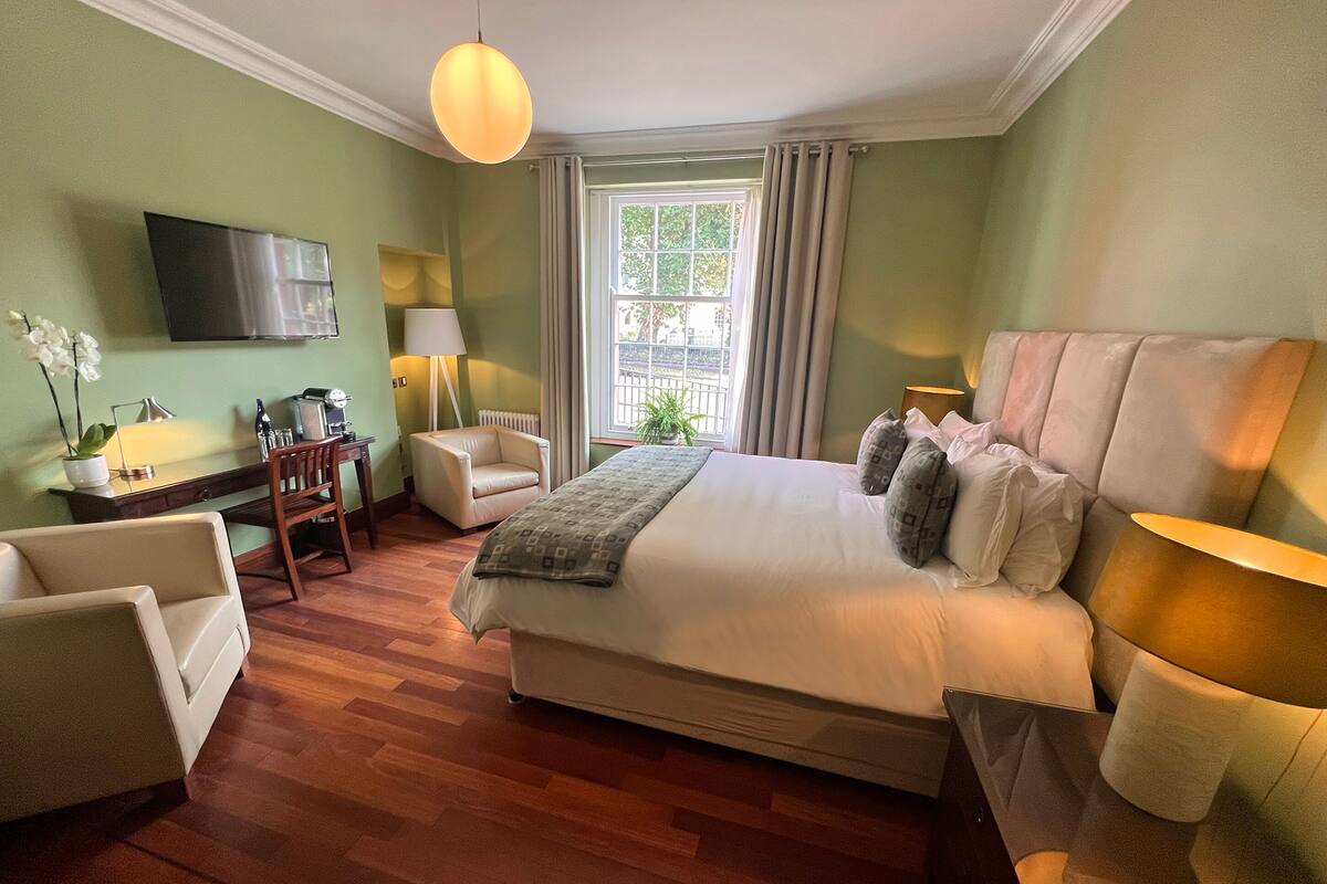A hotel room with a large bed, two armchairs, a desk with a lamp, a TV, and a window with curtains.
