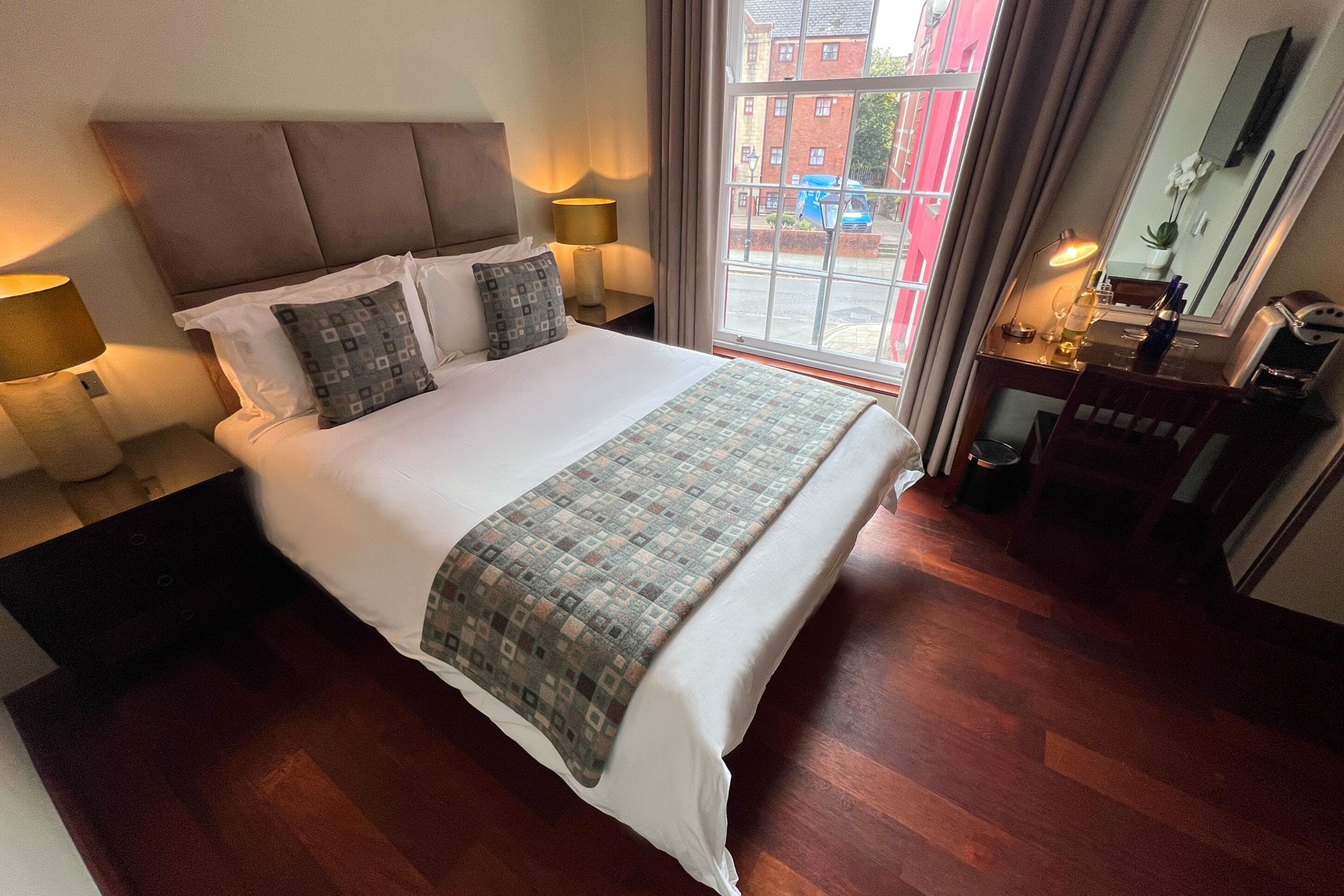 A hotel room with a large bed, two bedside lamps, a wooden floor, a window with a view of buildings, and a small desk with a mirror and a bottle.