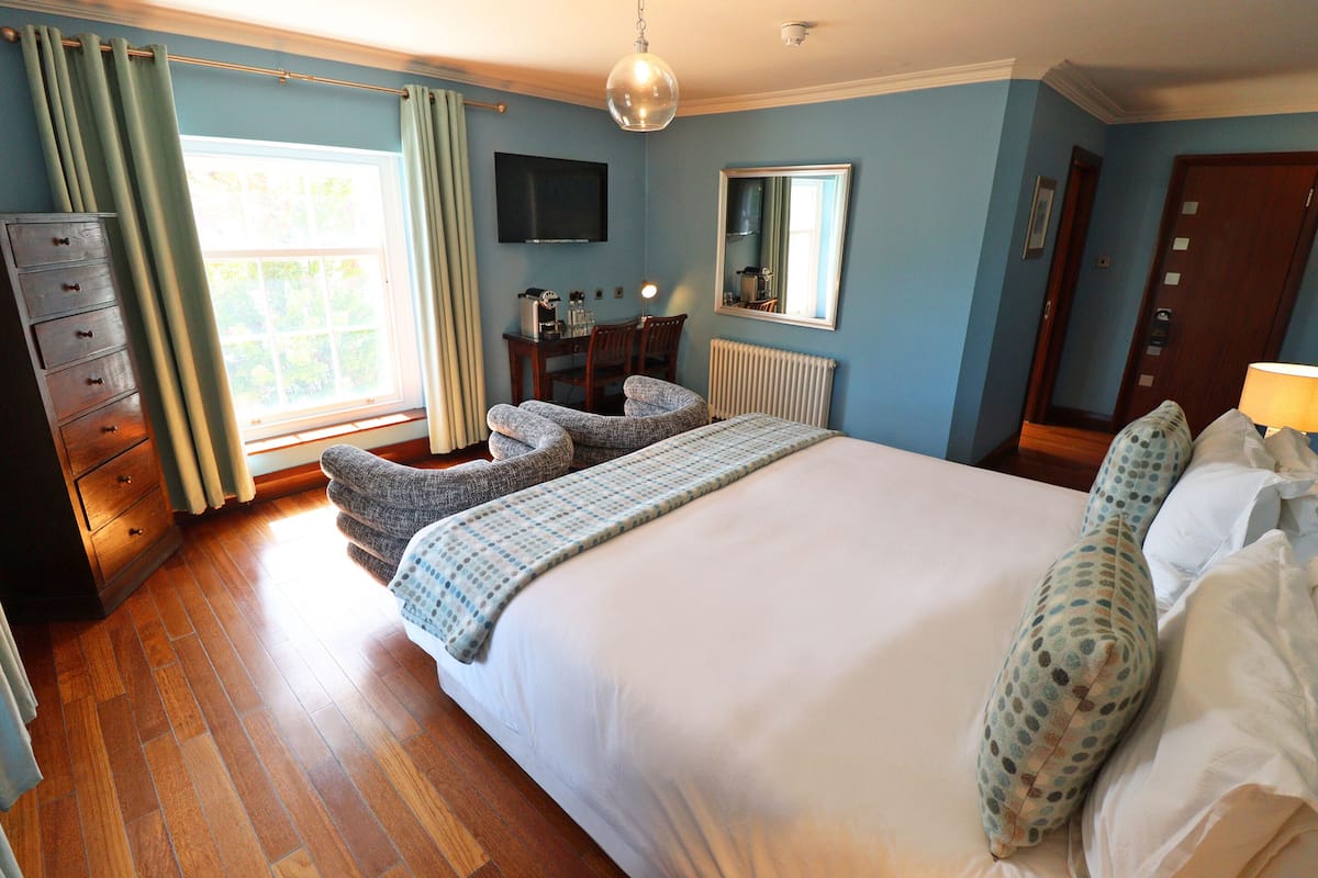 A bedroom with a large bed, a dresser, a chair, a desk with a chair, a television, a mirror, and a window with curtains.