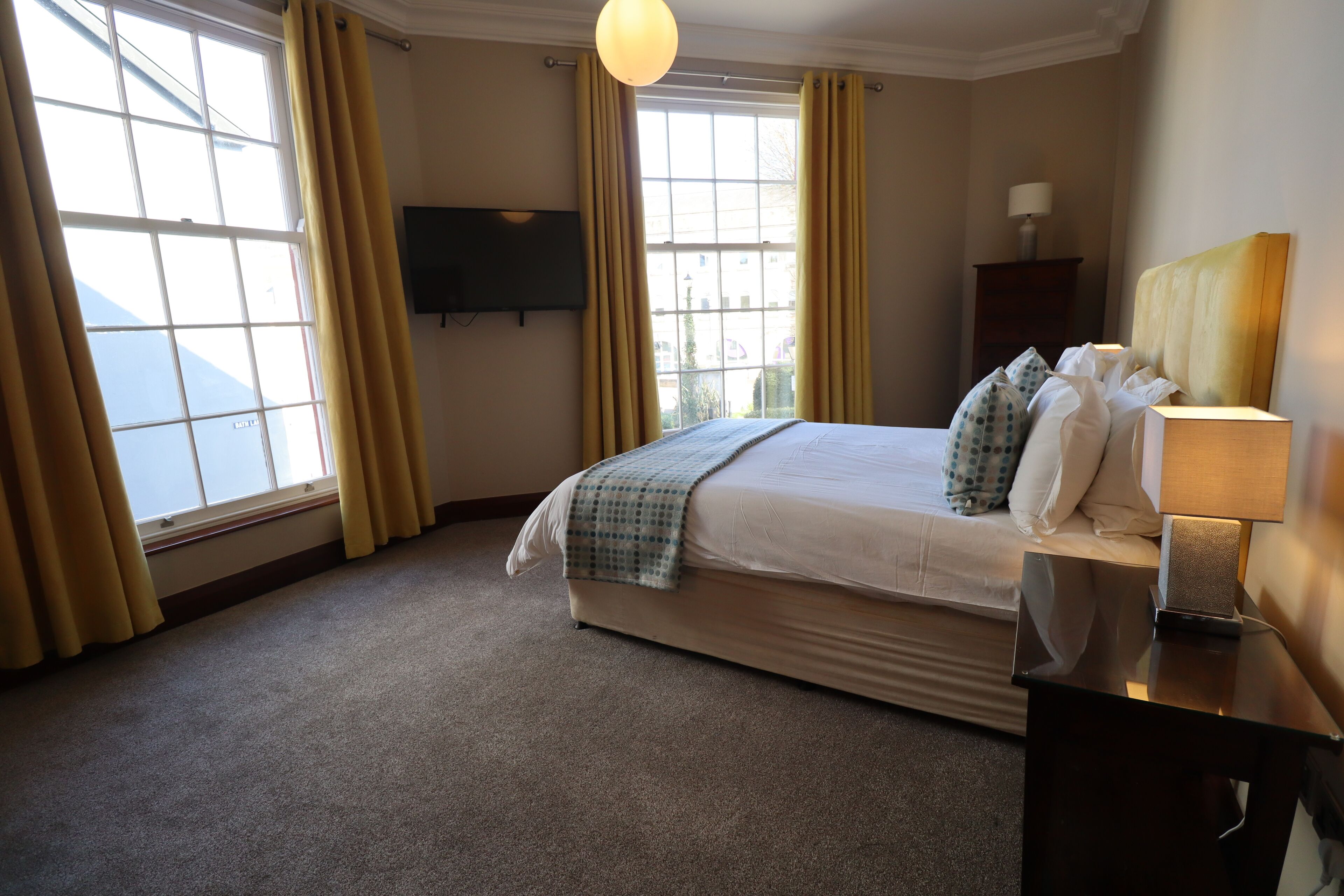 A hotel room with a large bed, a bedside table with a lamp, a TV mounted on the wall, and a window with curtains.