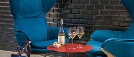 A modern lounge area with two blue armchairs, a small red table, a bottle of wine, two wine glasses, and a blue handbag.