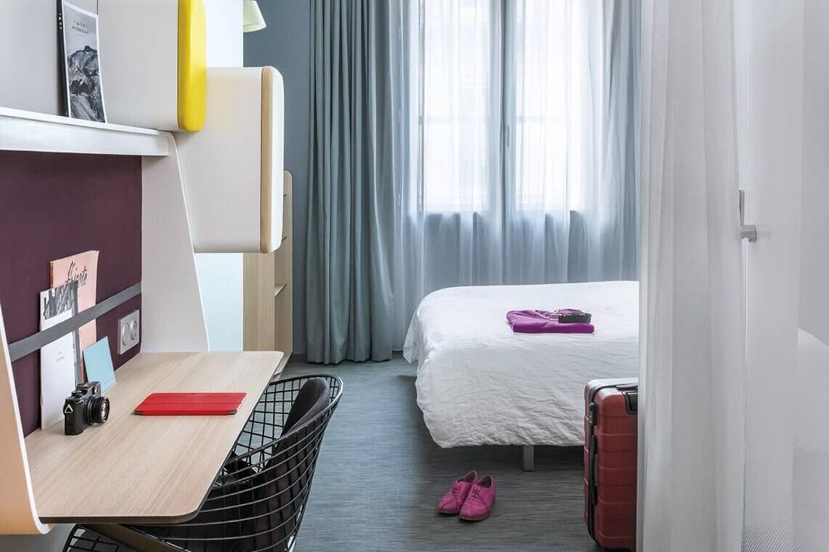 Une chambre d’hôtel avec un lit, un bureau, une chaise, une valise et une paire de chaussures.