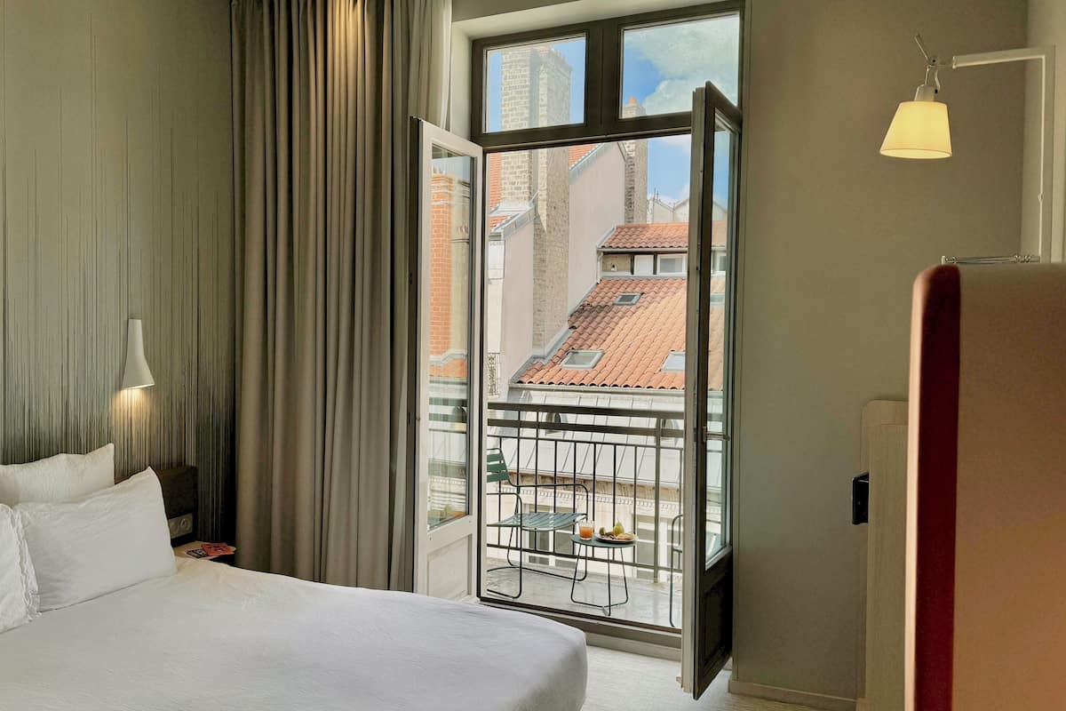 Une chambre d’hôtel avec un grand lit, une vue sur le paysage urbain par la fenêtre et un balcon équipé d’une table et de chaises.