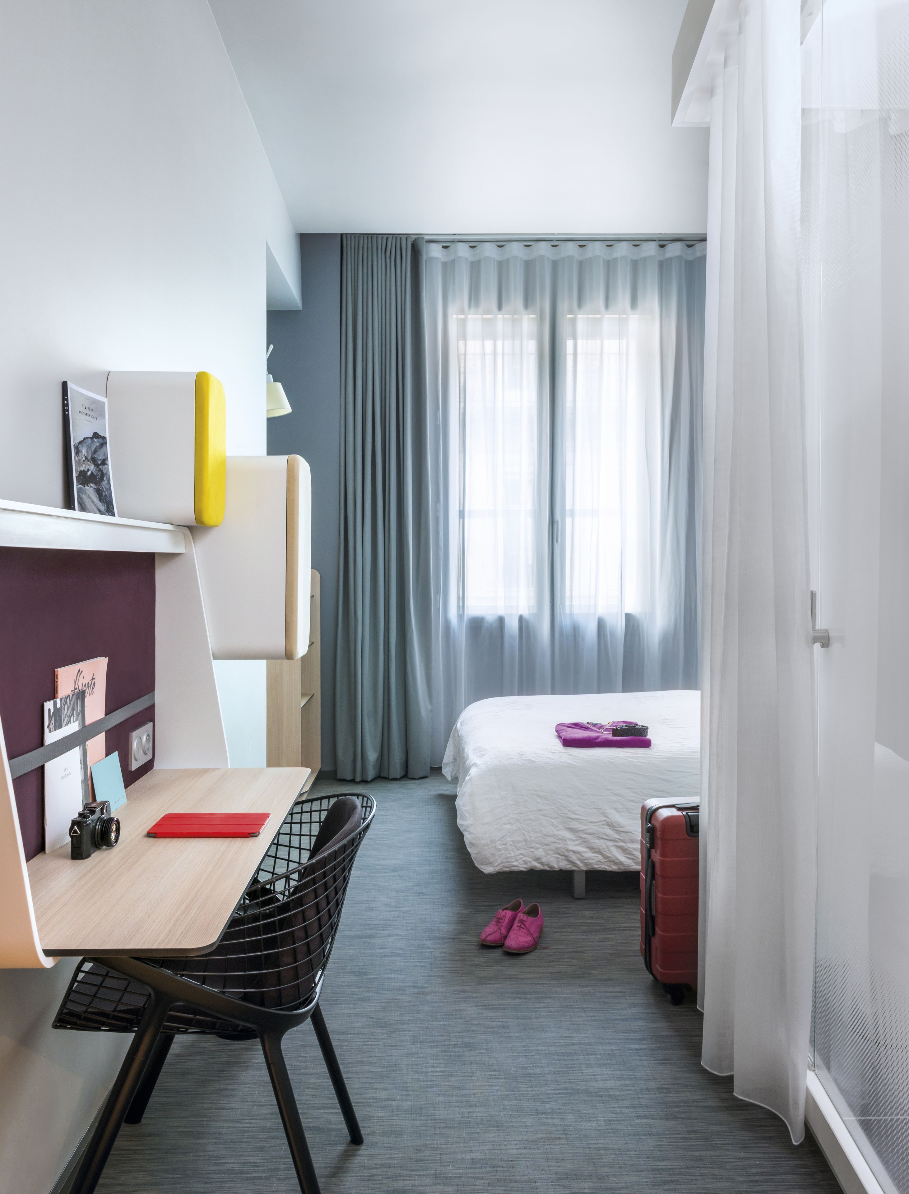 A compact hotel room with a desk, chair, bed, and a suitcase.