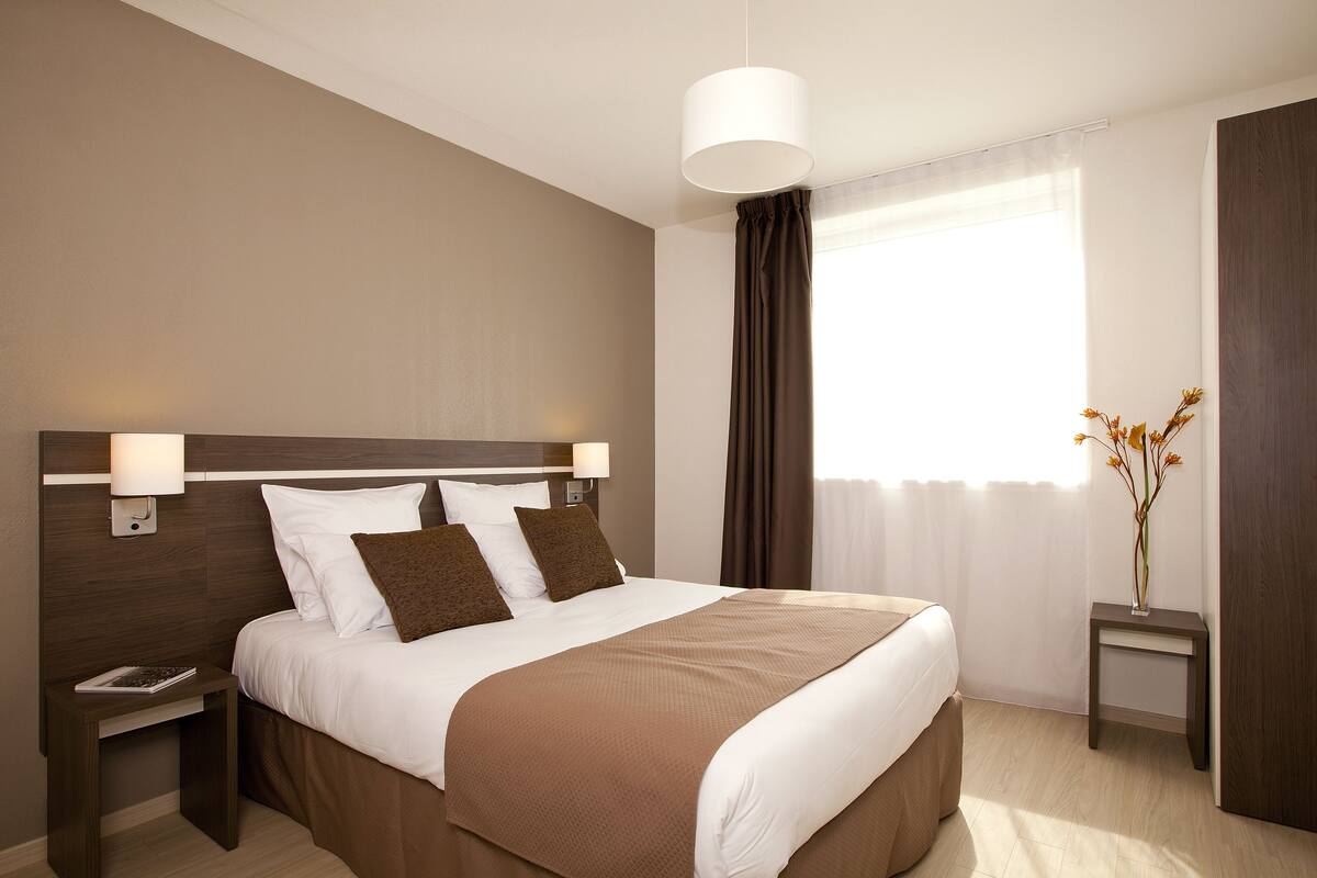 Une chambre d’hôtel avec un grand lit, une table de chevet, une lampe de chevet, une fenêtre avec des rideaux et un vase rempli de fleurs.