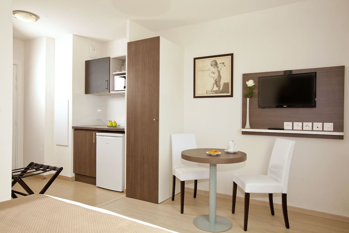 Une chambre d’hôtel compacte dotée d’une kitchenette, d’un coin repas avec une table ronde et deux chaises, d’un téléviseur à écran plat fixé au mur et d’un tableau encadré au-dessus du téléviseur.