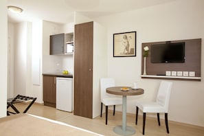 Habitación de hotel compacta con kitchenette, zona de comedor con una mesa redonda y dos sillas, televisor de pantalla plana montado en la pared y un cuadro enmarcado sobre el televisor.