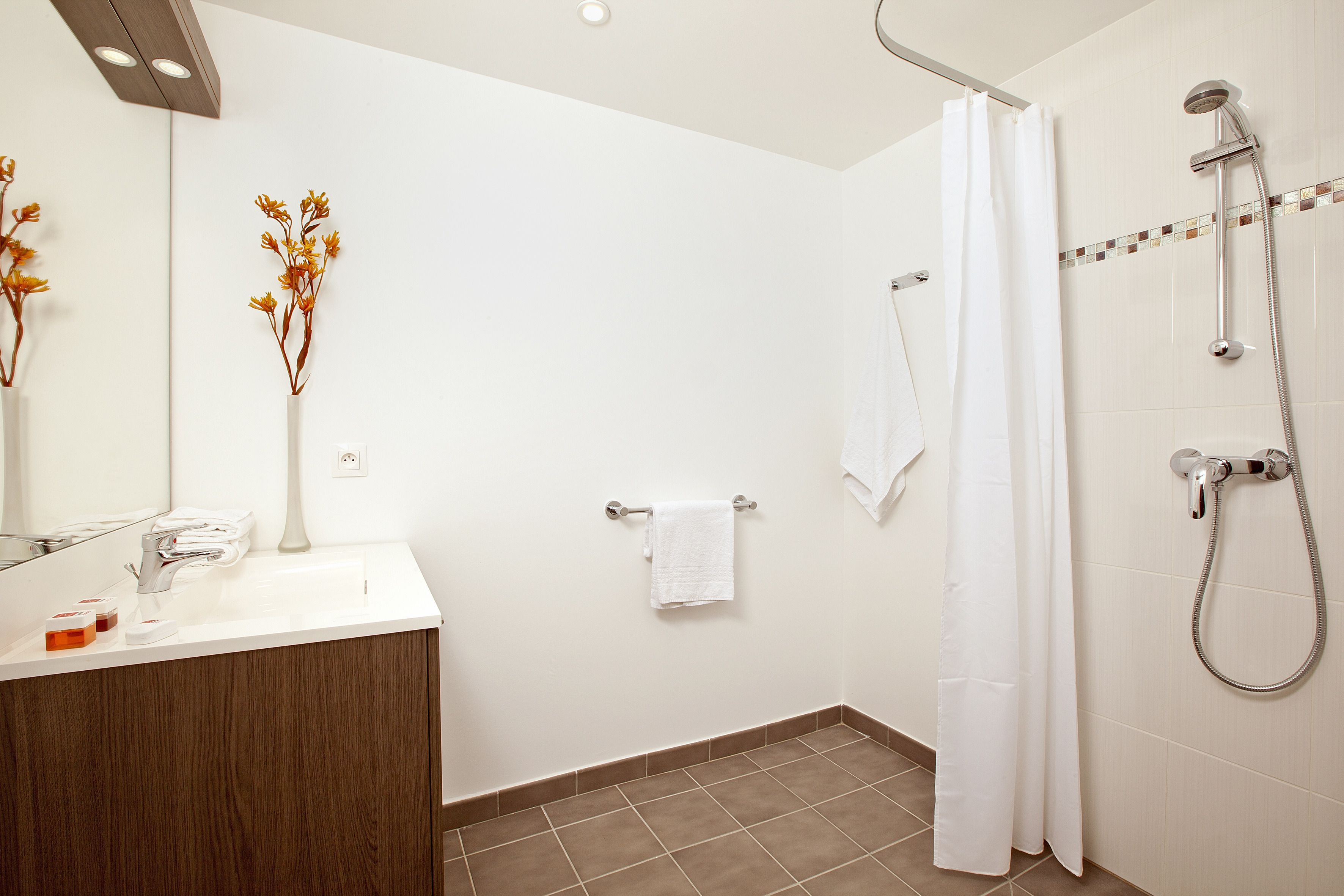 Une salle de bain moderne avec un lavabo blanc, un miroir, une douche équipée d’un pommeau de douche à main et un porte-serviettes.