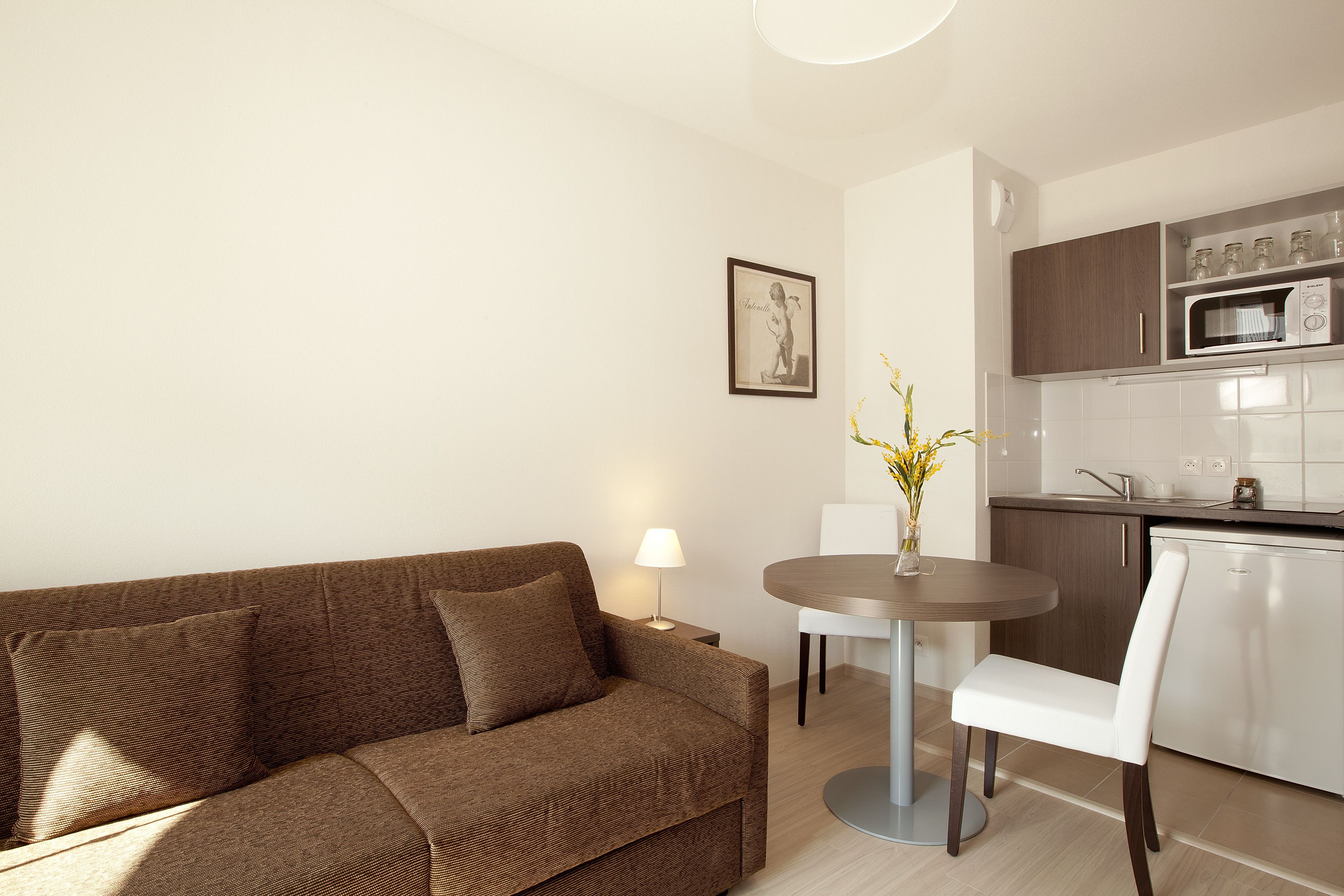 A modern living room with a brown sofa, a round dining table, and a white chair. A small table lamp is on the side table.