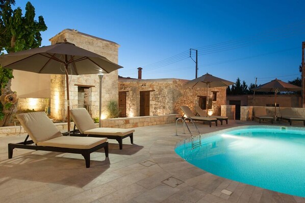 Un espace extérieur avec une piscine, des chaises longues et des parasols, des murs en pierre et un ciel dégagé.