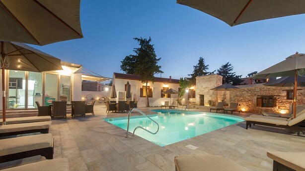 Zona de piscina exterior con sillas de descanso y sombrillas, rodeada de muros de piedra y un edificio con puertas de cristal.
