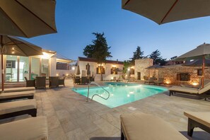 An outdoor swimming pool area with lounge chairs and umbrellas, surrounded by stone walls and a building with glass doors.