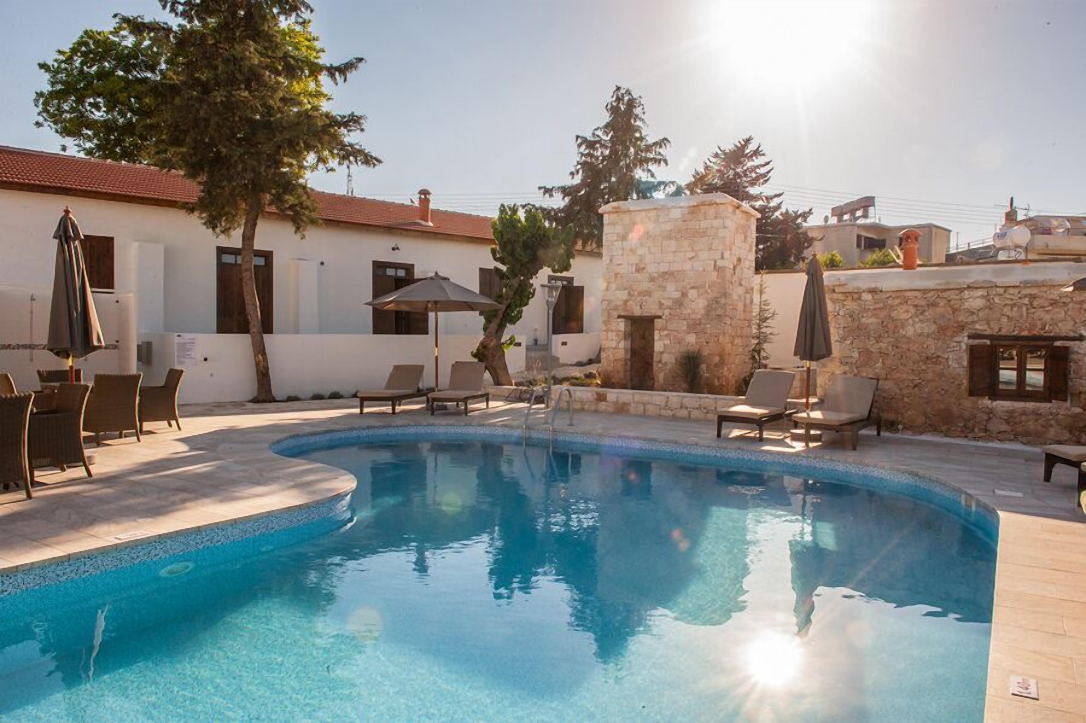 Une piscine entourée de chaises longues et de parasols, elle-même bordée de murs en pierre et d’un bâtiment blanc au toit rouge.