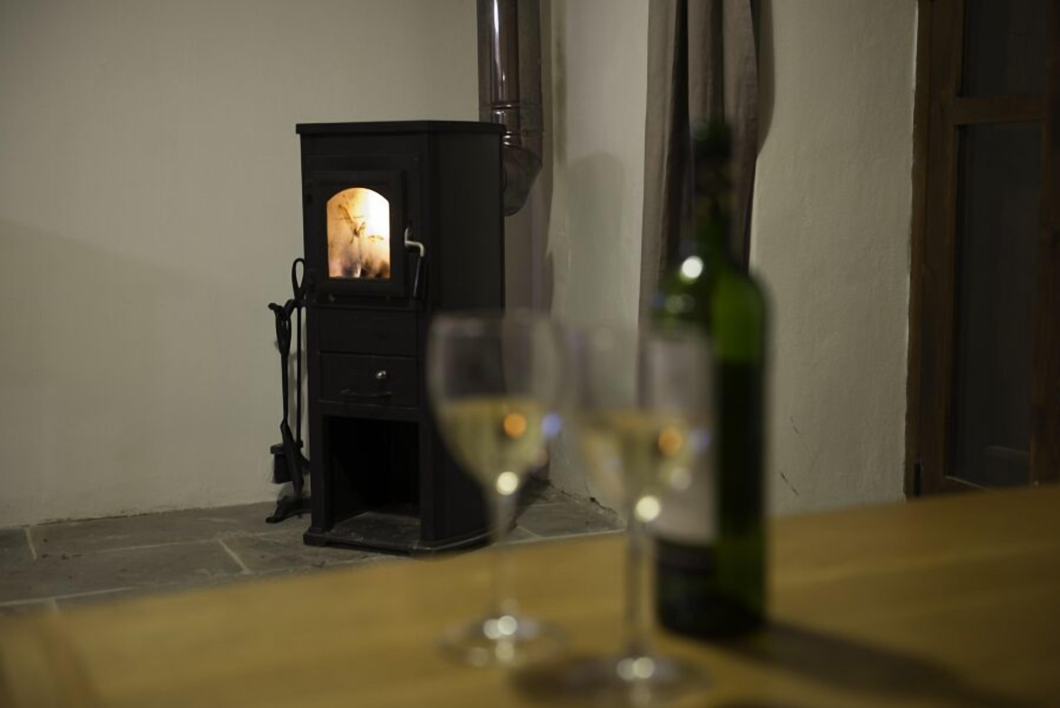 Una estufa de leña con puerta de vidrio, una botella de vino y dos copas sobre una mesa.