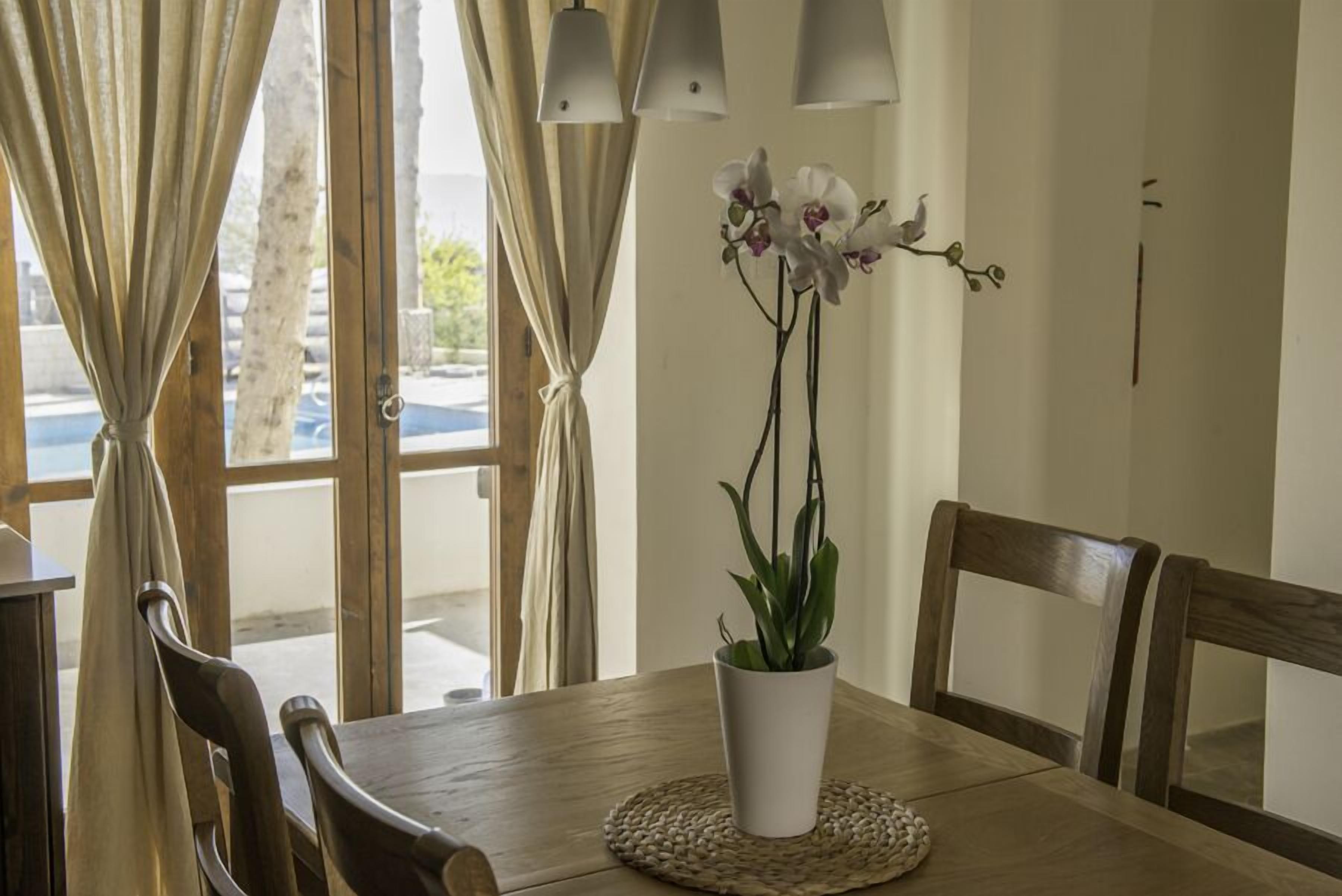 A wooden dining table with a white vase of orchids, wooden chairs, and a woven placemat.