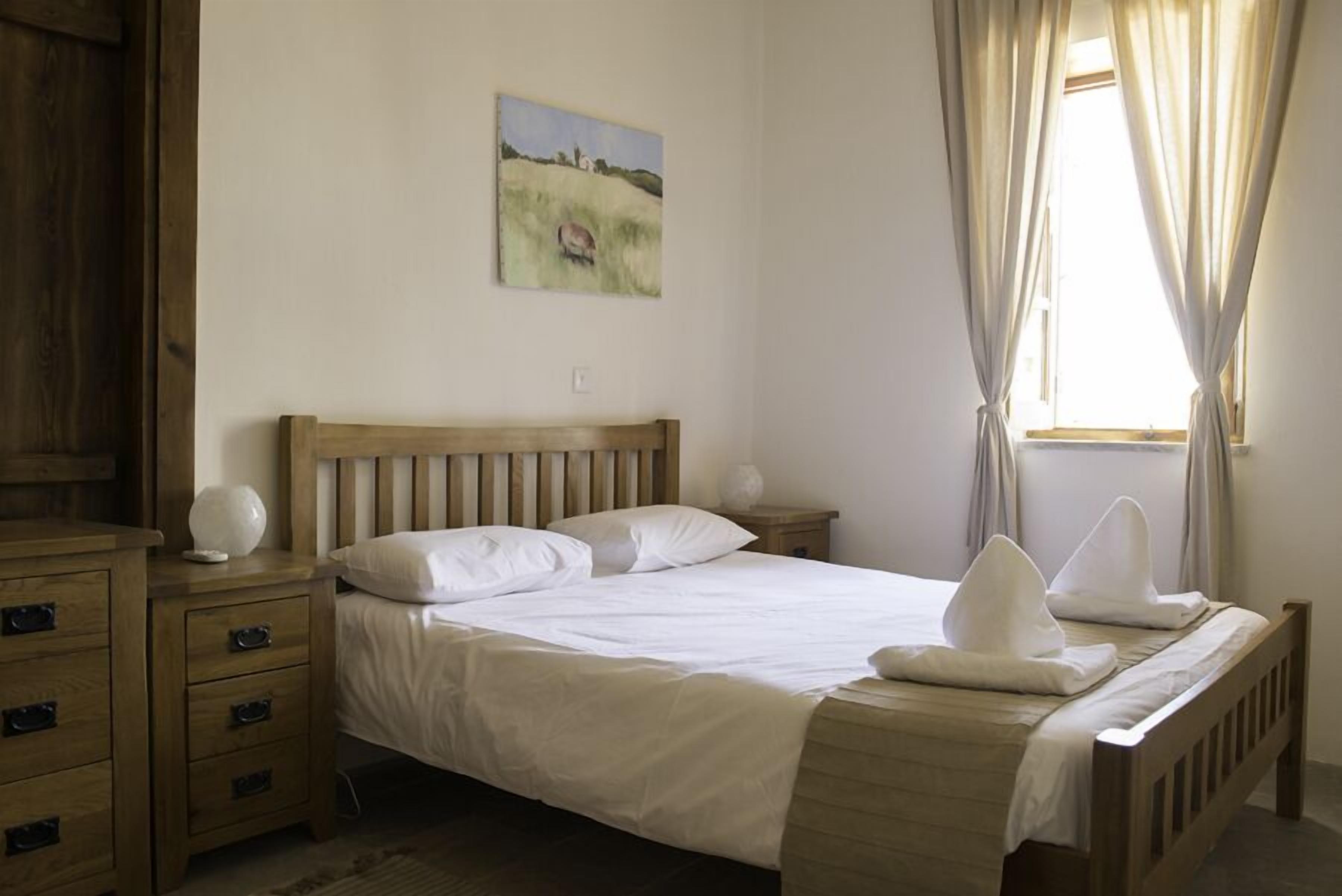 Un dormitorio con cama de madera, ropa de cama blanca, una mesita de noche con lámpara y un cuadro en la pared.