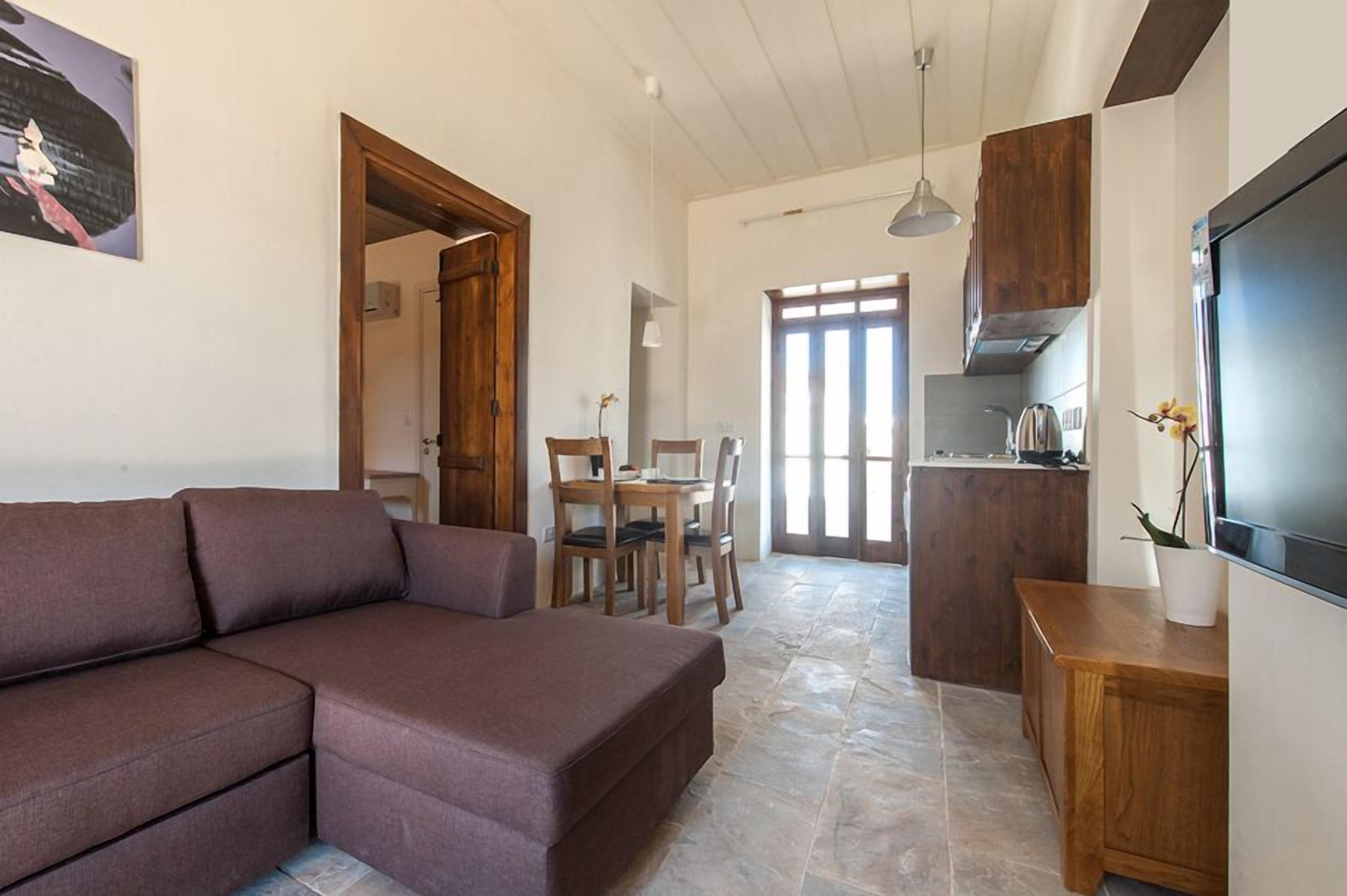 A living room with a brown sofa, a dining table with chairs, a kitchen area with a stove, and a TV on the wall.