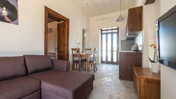 A living room with a brown sofa, a dining table with chairs, a kitchen area with a stove, and a TV on the wall.