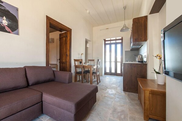 A living room with a brown sofa, a dining table with chairs, a kitchen area with a stove, and a TV on the wall. - Ktima 1937 Kannides (Droushia)
