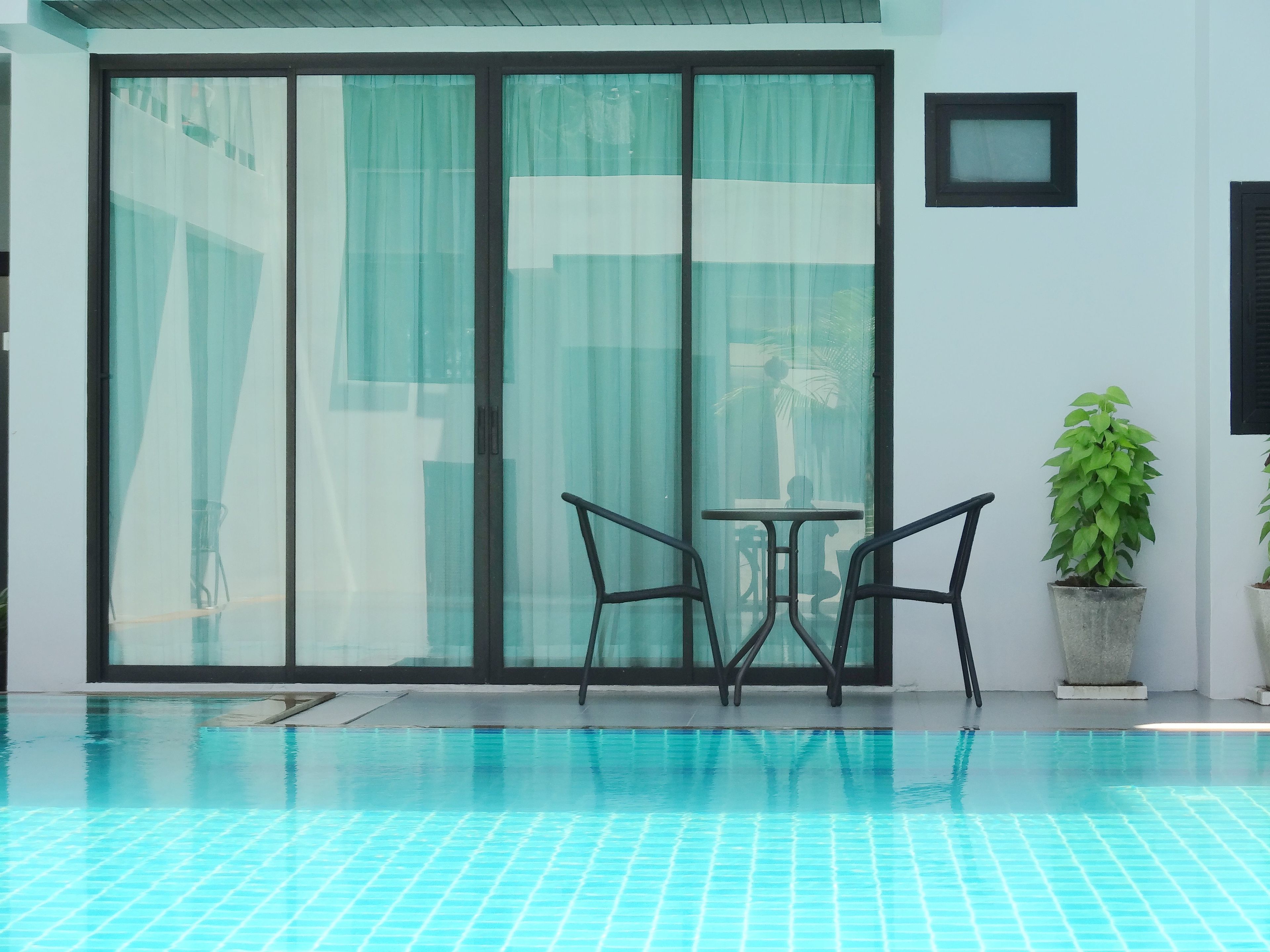 A modern indoor pool area with a glass partition, two chairs, a small table, and a potted plant.