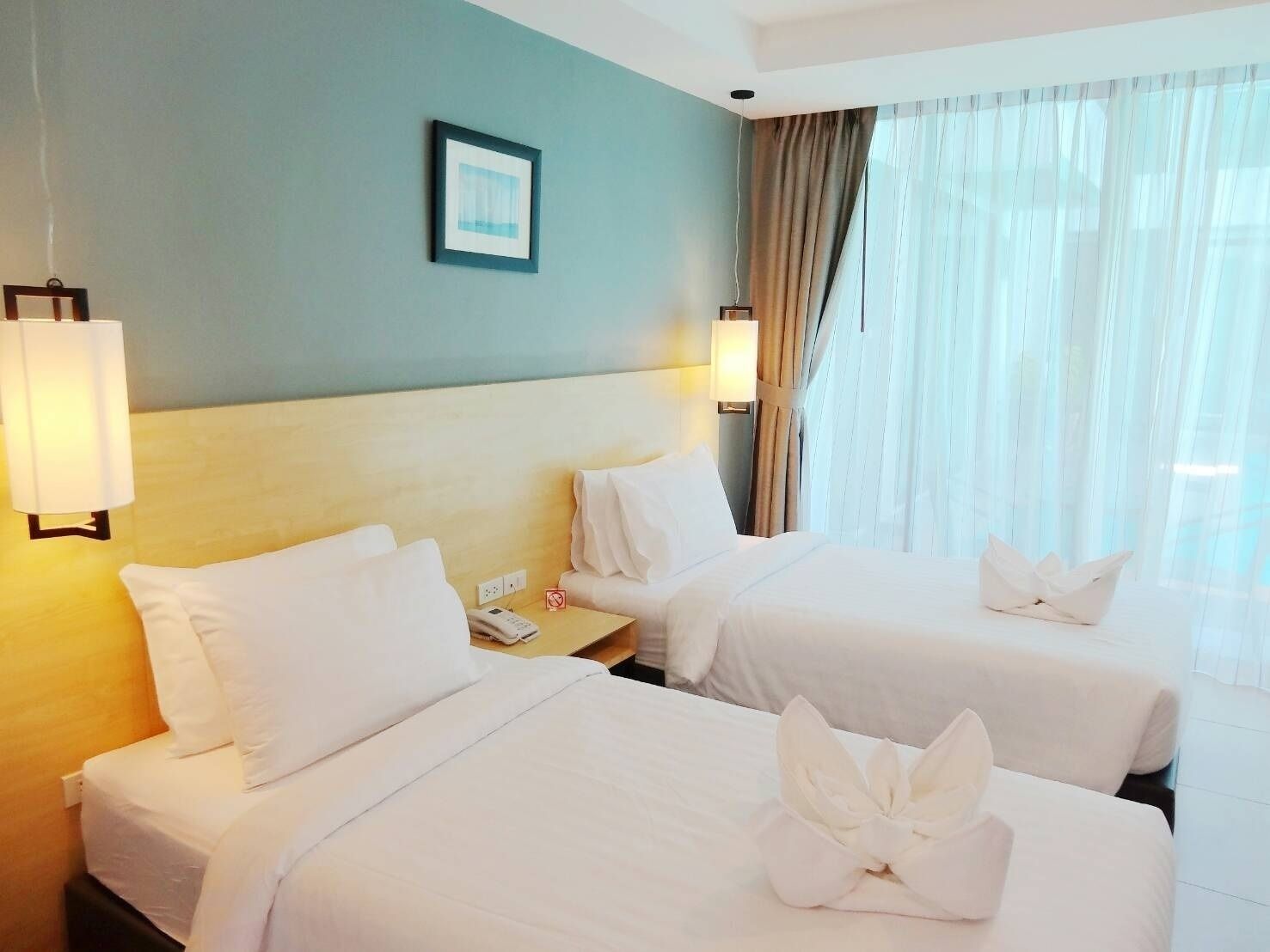 A hotel room with two beds, a headboard, a bedside table with a phone, a lamp, and a window with curtains.