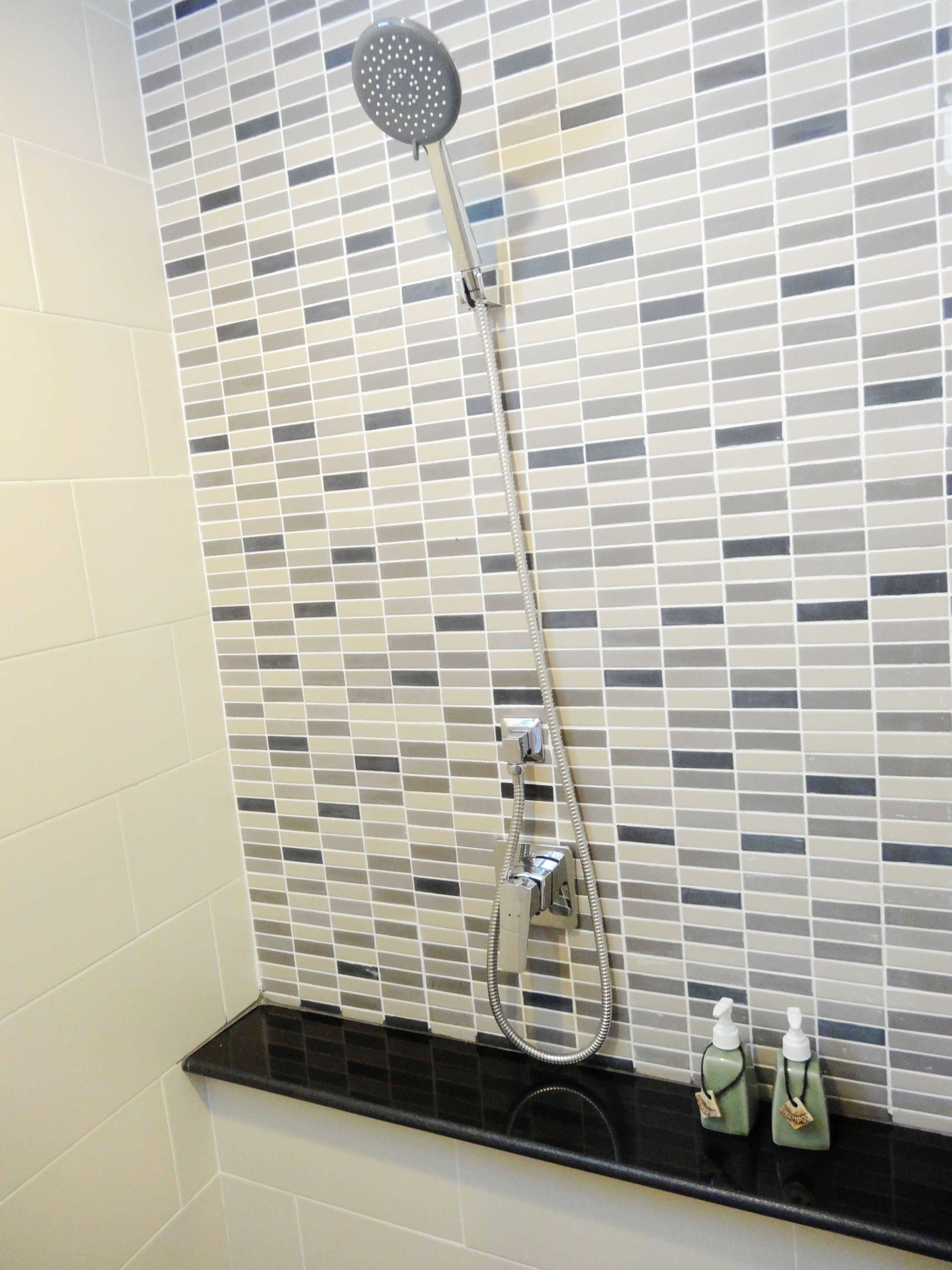A shower with a handheld showerhead and a wall-mounted control. There are two bottles of shampoo on a black countertop below the shower.