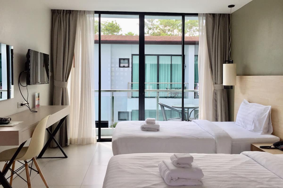 Habitación de hotel con dos camas, un escritorio, televisor y balcón con vistas a edificios.