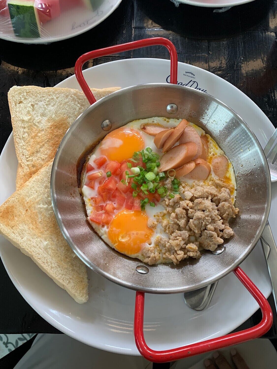 Un plat de petit-déjeuner composé d’œufs, de saucisses et de tomates servis sur des tranches de pain grillé, le tout présenté dans un plat en métal sur une assiette blanche à laquelle est fixée une anse rouge.