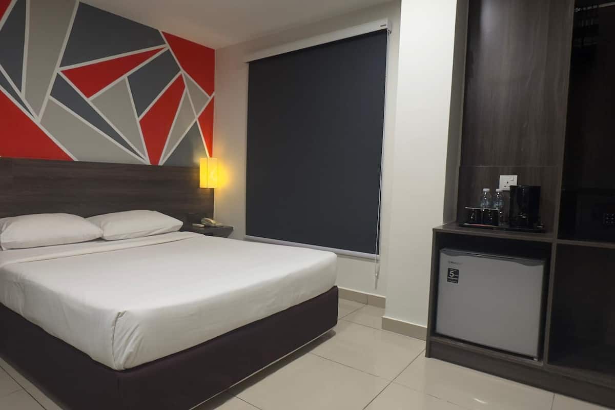 A modern hotel room with a large bed, a nightstand, a desk with a lamp, and a small refrigerator.
