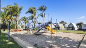 Un parque infantil con un tobogán y estructuras para escalar, rodeado de arena y palmeras.