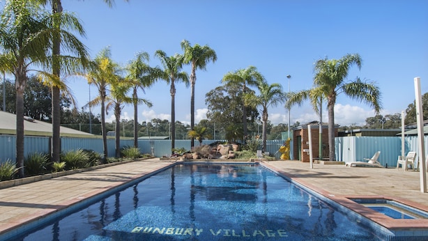 Una piscina con la inscripción "HUNBURY VILLAGE" en el agua, rodeada de palmeras y sillones de descanso.