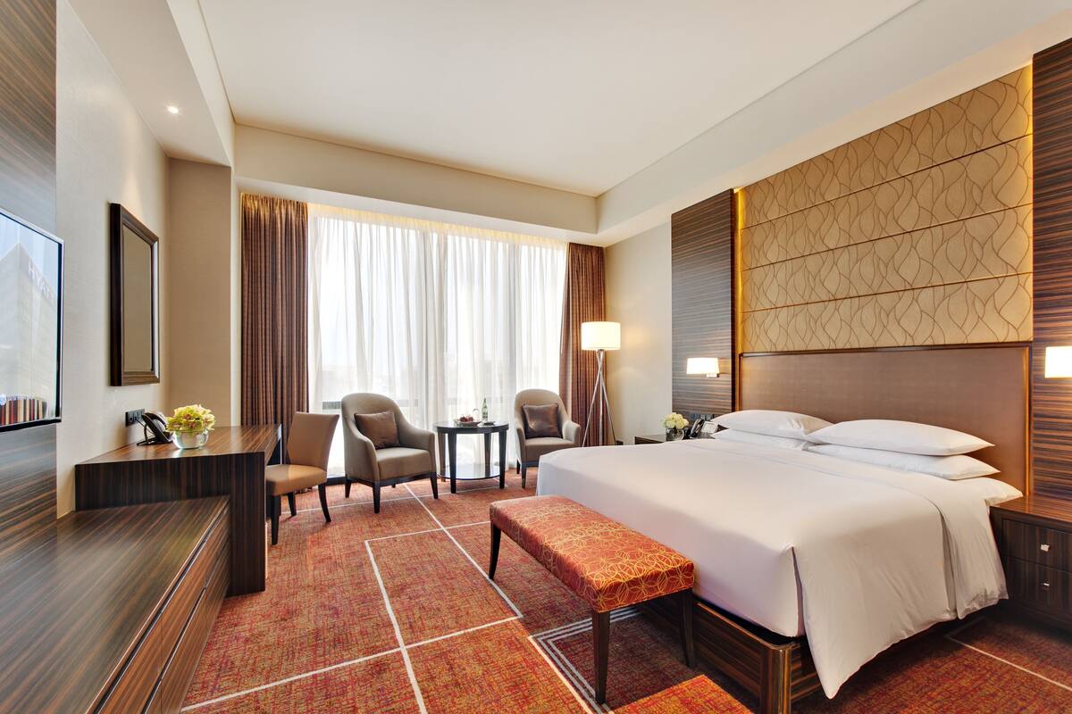 A hotel room with a large bed, a desk, two armchairs, a small table, and a bench.