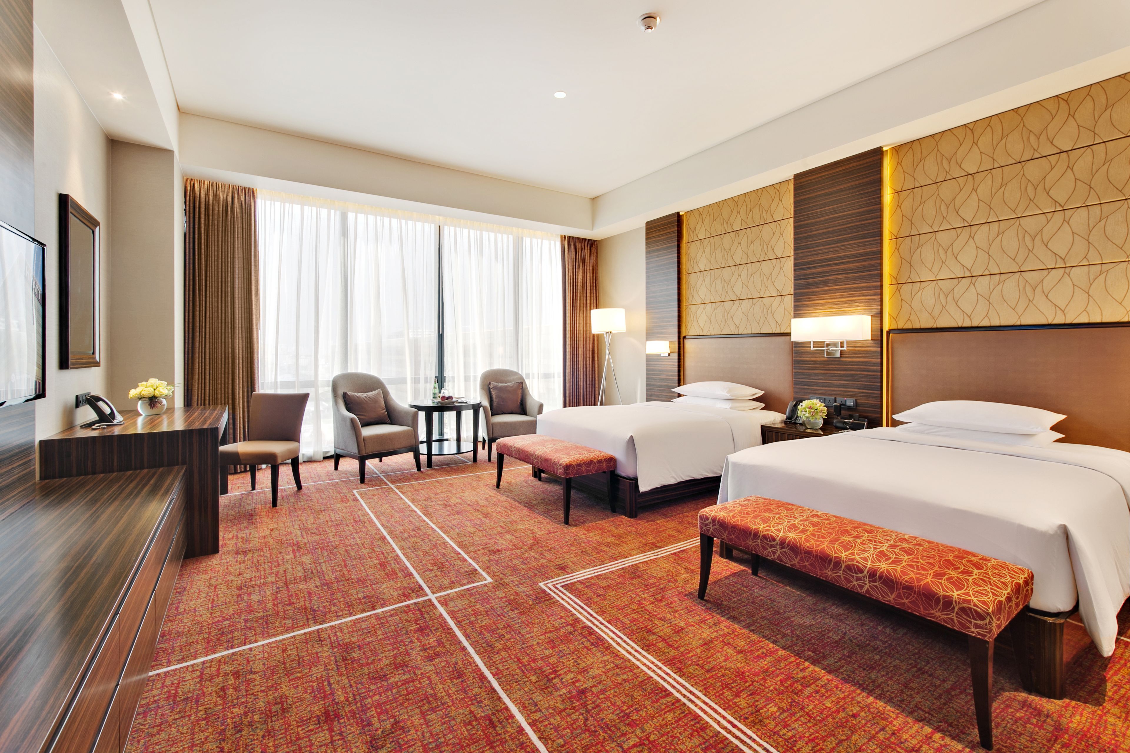 A hotel room with two beds, a desk, a chair, a bench, and a large window with curtains.