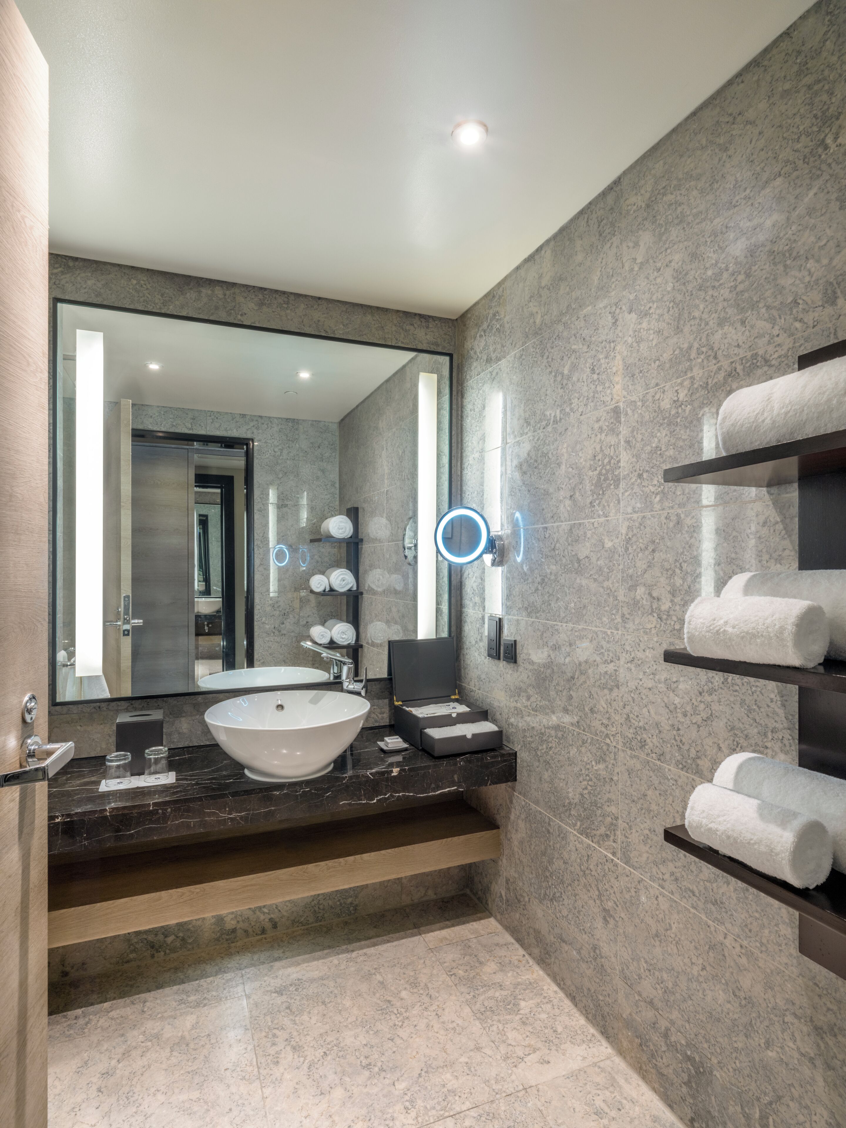 A modern bathroom with a large mirror, a round mirror, a white sink, and shelves with towels.
