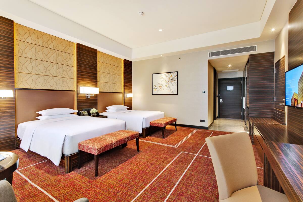 A hotel room with two beds, a bench, a TV, and a desk.
