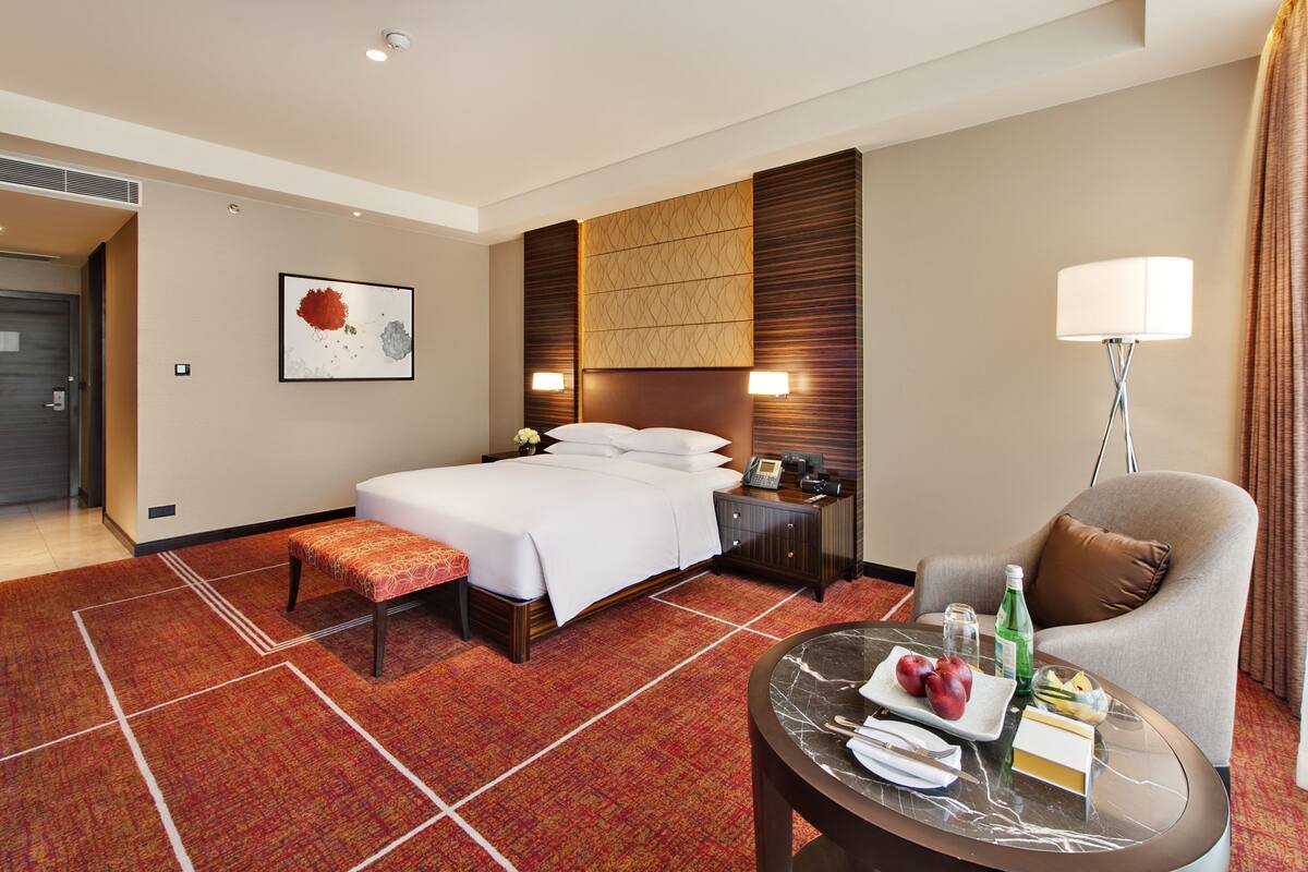 A hotel room with a large bed, a bedside table with a lamp, a chair, a small table with food and drinks, and a wall-mounted artwork.