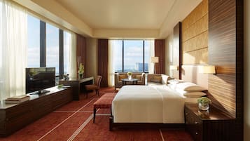 Habitación de hotel con una cama grande, un escritorio con televisor, una zona de estar con sofá y sillones, y vistas a la ciudad a través de amplias ventanas.