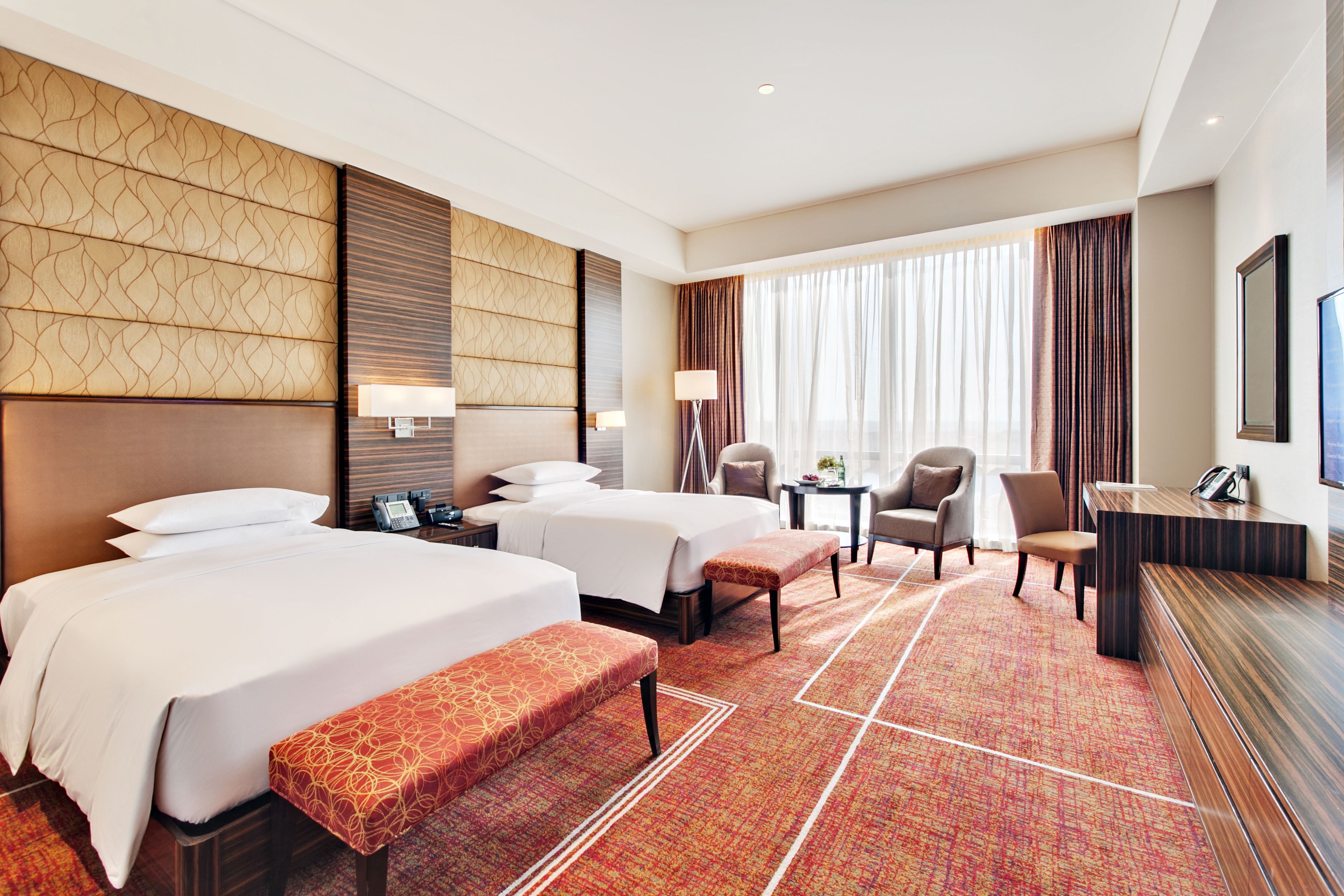 A hotel room with two beds, a desk, a chair, a bench, and a large window with curtains.