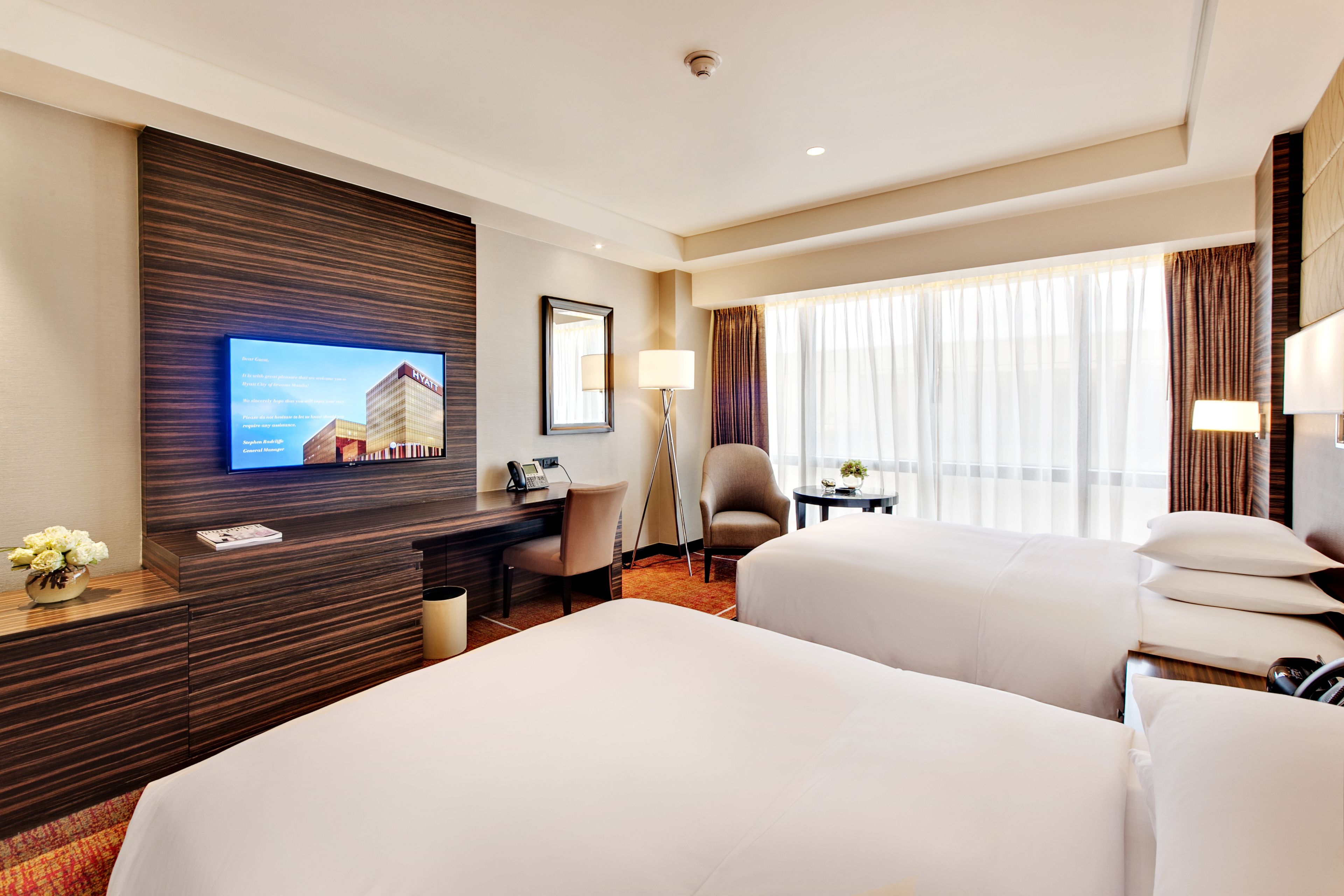 A hotel room with two beds, a desk, a chair, a TV mounted on the wall, and a window with curtains.