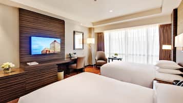 A hotel room with two beds, a desk, a chair, a TV mounted on the wall, and a window with curtains.