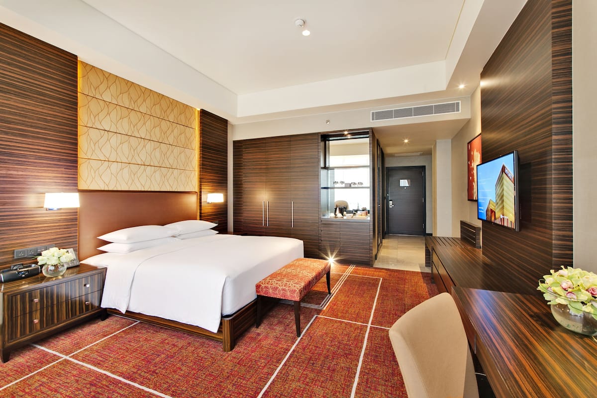 A modern hotel room with a large bed, a wooden headboard, a bedside table with a lamp, a TV, and a small bench.