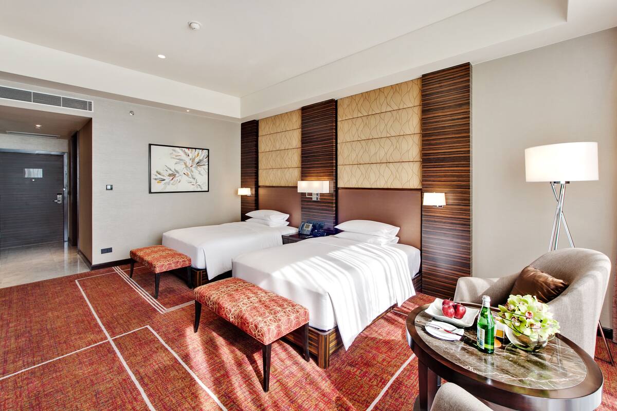 A hotel room with two beds, a sitting area with a table, and a view of the hallway.