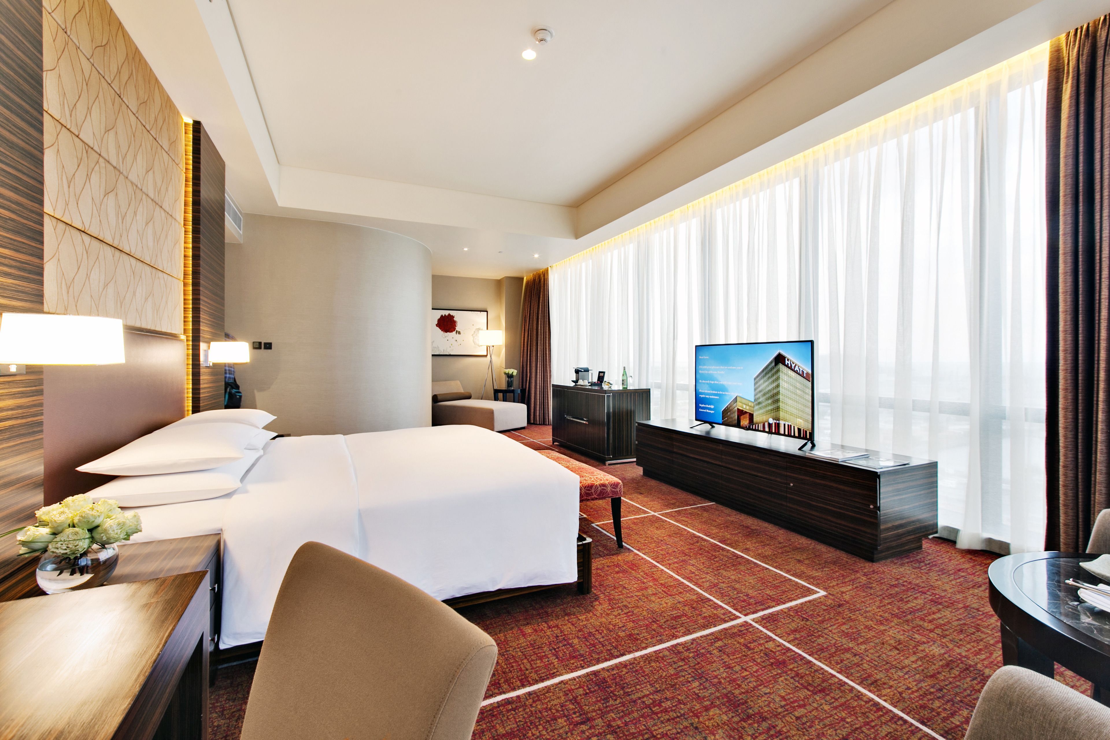 A hotel room with a large bed, a desk with a lamp, a television, and a view of a building through the window.