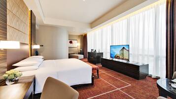 A hotel room with a large bed, a desk with a lamp, a television, and a view of a building through the window.