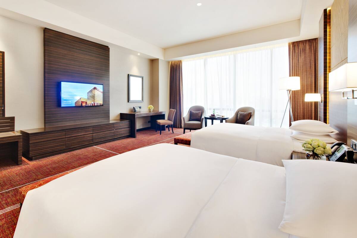 A hotel room with two beds, a desk, a TV, and a large window with curtains.