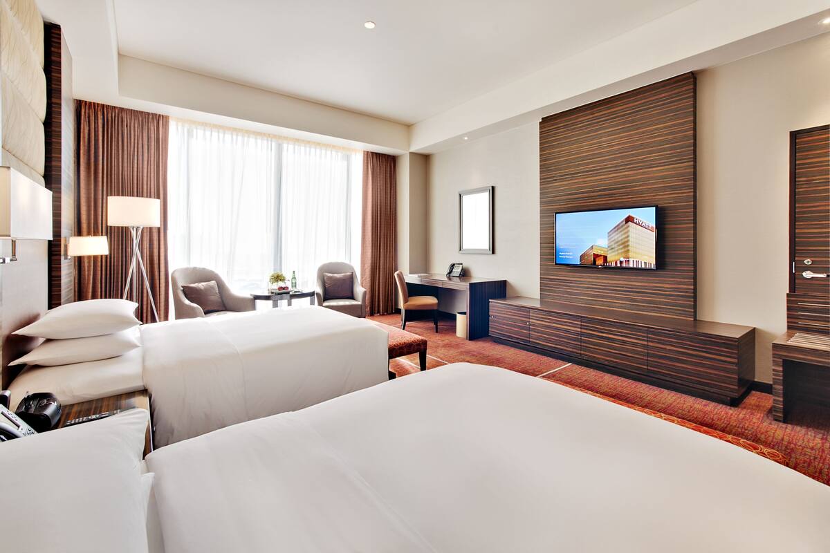 A hotel room with two beds, a desk, a chair, a TV, and a large window with curtains.